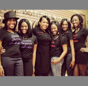 Five smiling women posing together indoors, dressed stylishly.